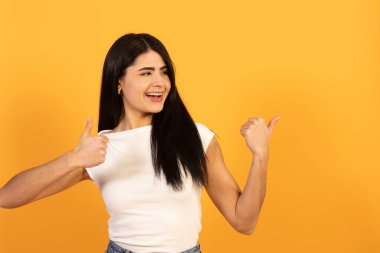 Uzun siyah saçlı mutlu bir genç kadın, geniş bir gülümseme, bir çift baş parmak yukarı jest sunuyor ve canlı sarı bir arka plan, başarı ve mükemmellik kavramları ileten