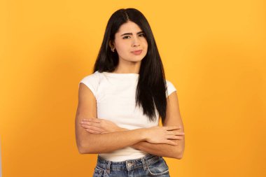 Kolları çapraz duran genç bir kadın, şüpheli bir yüz ifadesi sergiliyor ve doğrudan kameraya bakıyor. Parlak sarı bir arka planda, şüphe ve hoşnutsuzluğu iletiyor.