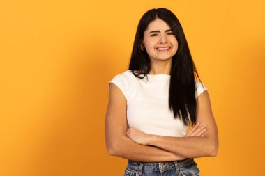 Kollarını kavuşturmuş, güven içinde kameraya gülümseyen genç bir kadın canlı sarı bir zemine karşı mutluluk, başarı ve olumlu duygular betimliyor.