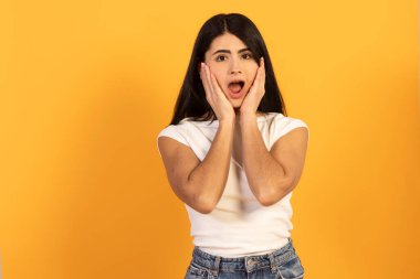 Uzun siyah saçlı genç bir kadın elleriyle yüzünü kapatıyor, canlı sarı bir stüdyo arka planında duygusal bir şok, sürpriz ya da inançsızlık ifadesi sergiliyor, doğrudan izleyiciye bakıyor.