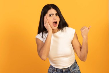 Şok olmuş, şaşırmış bir ifadeye sahip genç bir kadın, yanak yanağa ve başparmağıyla yan tarafı işaret ediyor, parlak sarı stüdyo geçmişine karşı beklenmedik haberlere tepki veriyor.