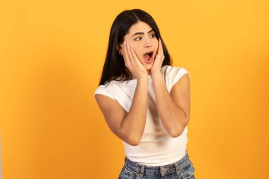 Beyaz tişörtlü, koyu renk saçlı genç bir kadın, yanaklarına el ele tutuşuyor, gözleri açık ve ağzı açık, şaşkın bir şekilde, canlı sarı bir arka plana karşı şaşkın, şaşkın bir tepki veriyor.