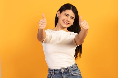 Gülümseyen genç bir kadın, parlak sarı bir arka plana karşı onay, başarı ya da mükemmel bir hizmet, somut pozitiflik ve teşvikle iki baş parmak hareketi gösterir.
