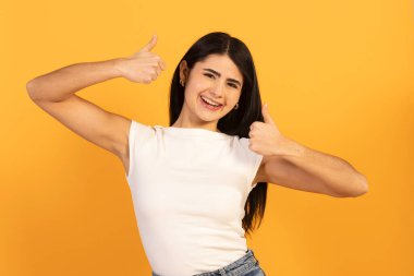 Uzun siyah saçlı, beyaz tişörtlü, neşeli bir şekilde gülümseyen genç ve mutlu bir kadın. Parlak sarı bir arka planda onay, anlaşma ve başarıyı ifade ediyor.