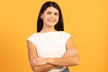 Genç esmer kadın kendine güvenen bir gülümseme ve çapraz kollarla poz veriyor, canlı sarı bir arka plana karşı duruyor, pozitifliğini ve kendine güvenini dile getiriyor.