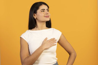 Sarı arka planda duran genç bir kadın, elini kalbinin üzerine koyup gülümseyerek samimiyet, minnettarlık, gurur ve dürüstlük ve samimi duygular ifade ediyor.