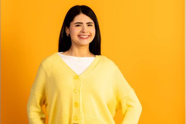 Uzun siyah saçlı, geniş gülümseyen, beyaz bluzunun üzerine sarı bir hırka giyen, kendine güvenen ve stüdyo ortamında neşeli, pozitif bir ruh hali sergileyen genç bir kadın.