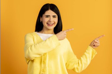 Mutlu genç bir kadın gülümsüyor ve iki elini de iki tarafa doğru gösteriyor, seçenekler sunuyor, ya da parlak sarı arka planda fotokopi alanı ile neşeli bir terfi veriyor.