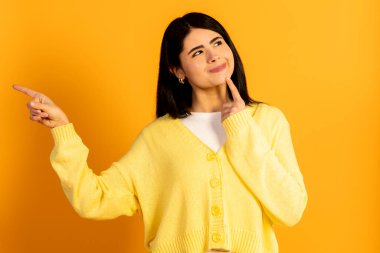 Bir karar vermeyi düşünen genç bir kadın, parmağını sol tarafı işaret ederek, seçenekleri ya da yönü belirterek, parlak sarı bir arkaplanda izole edilmiş.