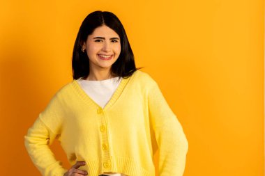 Sarı hırkalı, koyu renk saçlı genç bir kadın canlı sarı bir arka plana karşı güler, neşeli, doğal iyimserlik ve modern gündelik tarz yayar.