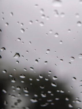 Cam bir pencere yüzeyine yapışmış yağmur damlaları yumuşak yansımaları ve bulanık gri arkaplanı olan doğal bir soyut desen yaratarak tazeliği ve barışçıl bir atmosferi sembolize eder.