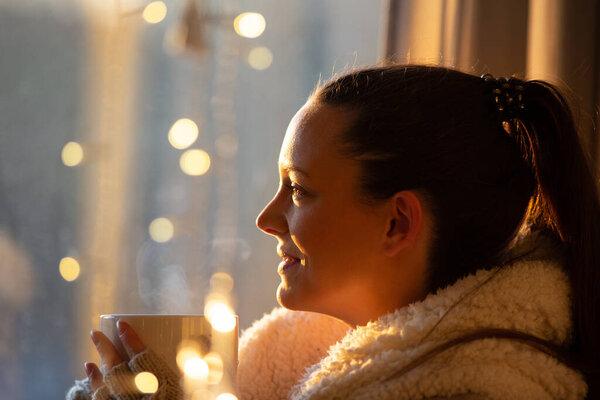 Beautiful girl covered with blanket holding cup of hot coffee with christmas lights in background, Festive moments concept