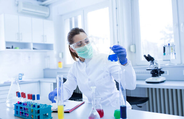 Молодой биолог в защитной маске, перчатках и очках тестирует некоторые химические вещества в бутылках в лаборатории