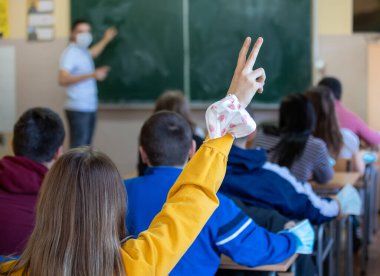 Sınıfta güvenlik maskesi takan, arka planda öğretmeniyle el kaldırmış liseli kızın dikiz görüntüsü. Eğitimde pandemik koruma kavramı