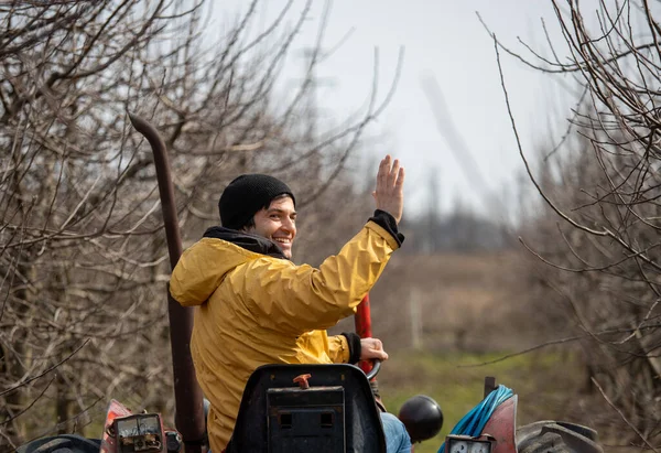 Rear view of farmer with raised hand for greeting driving tractor in orchard in winter time