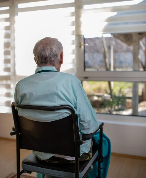 Rear view of senior man in pajamas sitting in wheelchair and looking through window in hospital