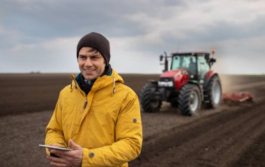 Tabletli yakışıklı bir çiftçinin portresi traktörün önünde duruyor arka plandaki tarlada yıpratıcı.