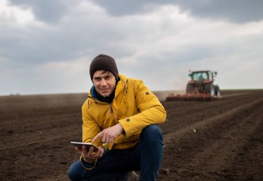 Tabletli yakışıklı bir çiftçinin portresi traktörün önüne çömelmiş arka plandaki tarlaya zarar veriyor.