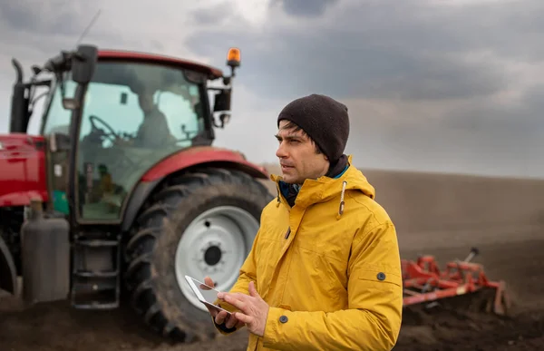 Portrait of handsome farmer with tablet standing in front of tractor harrowing in field in background