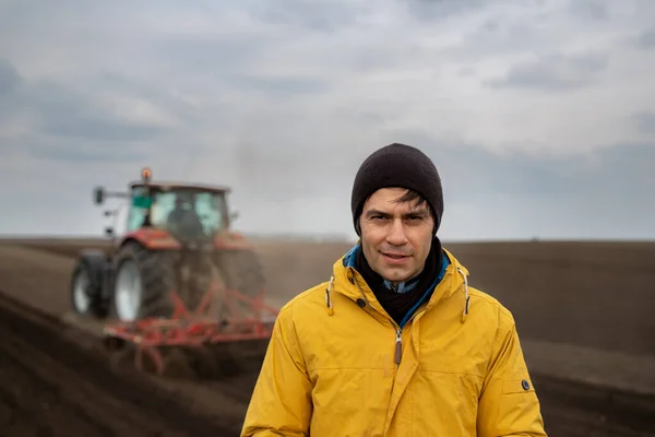 Portrait of handsome farmer standing in front of tractor harrowing in field in background