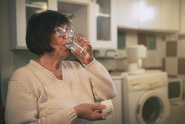 Mutfakta oturan, hap ve su içen yaşlı bir kadın.