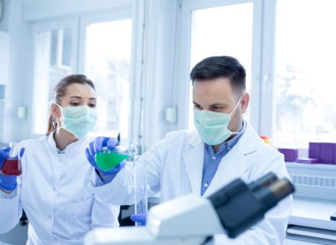 Erlenmeyer şişesini tutan deneyleri araştıran genç bilim adamları. Laboratuvarda kadın ve erkek teknisyen yüz maskesi takan sıvı ölçümleri yapıyor..