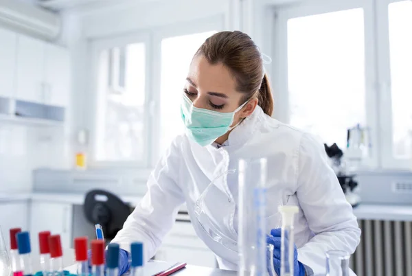 实验室技术人员使用药瓶监测写作实验 年轻的女科学家戴着口罩在实验室做医学研究 — 图库照片