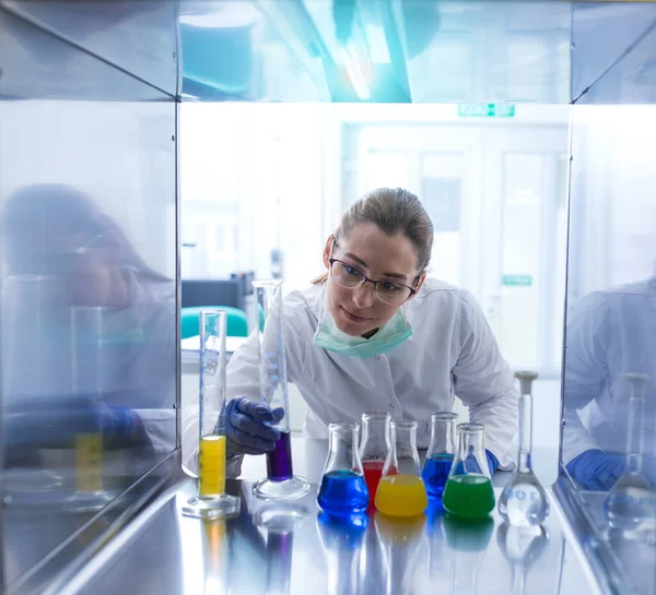 研究室で働く女性は研究実験を行う 顔マスク手袋エルレンマイヤーフラスコとシリンダー内の液体を監視する若い科学者研究室の技術者 — ストック写真