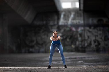 Genç bir kadın öne eğilip esneme hareketi yapıyor. Kentsel alanda egzersiz için ısınan sporcu gülümsüyor. 