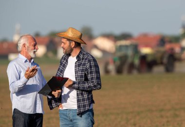 Çiftçi ve sigorta temsilcisi konuşuyor, tartışıyorlar. İki adam traktör çiftçilik makinesinin önünde pano ile pazarlık yapıyor.