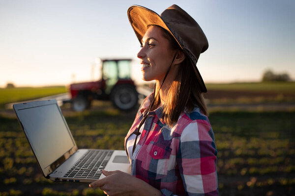 Young agronomist using computer innovation in food production. Female farmer standing in field with tractor machinery in background. 