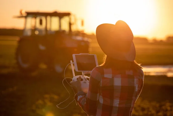 フィールドの農業でドローンコントローラの技術革新を使用して若い農家 夕方にトラクター機械を背景に現場に立つ女性農学者 — ストック写真
