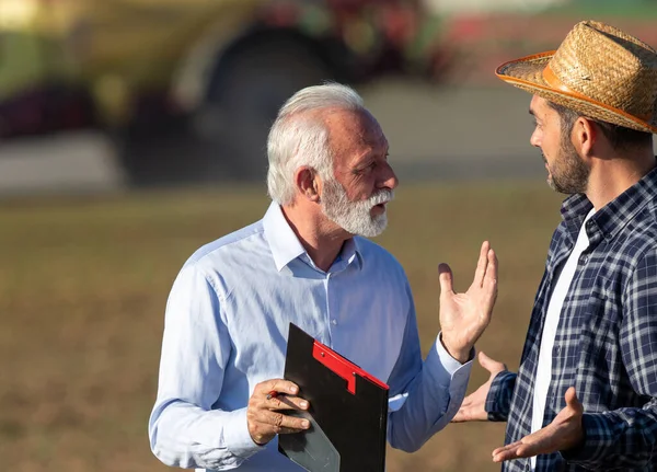 2人の男が機械を背景に現場で説明することについて話していた 農家保険販売代理店代表クリップボードを保持交渉 — ストック写真