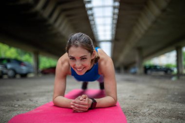 Atletik kız gündüz vakti otoparkta egzersiz yapıyor. Sportif genç bir kadın pembe hasırın üzerinde tahtasını yapıyor ileriye bakıyor gülümsüyor. 