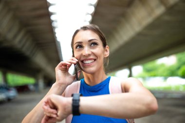 Gülümseyen genç kadın egzersiz sonrası cep telefonu ve akıllı saat kullanıyor. Köprünün altındaki otoparkta duran sporcunun portresi.
