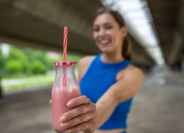Athletic woman in sportswear in background smiling holding milkshake. Close up of strawberry smoothie in glass jar bottle in focus.