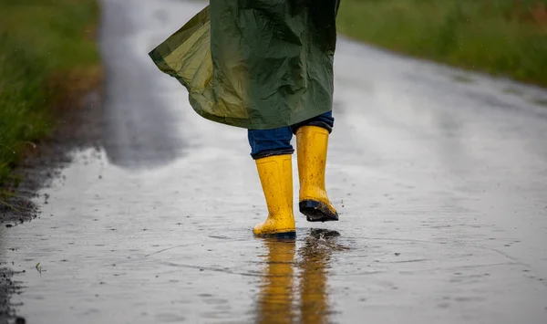 Close up of farmer\'s legs in yellow gumboots and green raincoat walking on puddles after rain in field