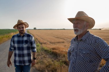 Young male farmer wearing plaid shirt and hat looking at camera. Elderly male farmer standing in field with hands on hips looking to side. 