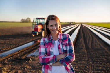 Female farmer standing with arms crossed looking at camera. Young farmer standing in field with plant foil and tractor smiling.