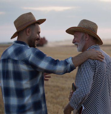 Two farmers wearing plaid shirts and straw hats talking. Young agronomist with hand of shoulder of old farmer laughing in wheat field during harvest in field in summer. 