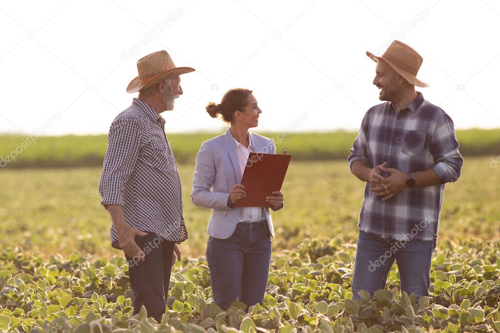 Young female agronomist holding clipboard smiling. Two male farmers negotiating with insurance sales rep standing in field.