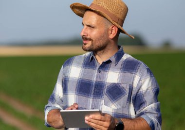 Tarımda modern teknolojiyi kullanarak sahada duran çekici genç tarımcı. Tablet tutan hasır şapkalı bir erkek çiftçinin portresi.