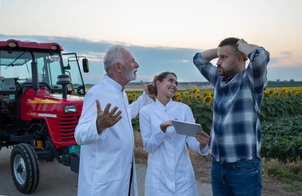 夏にトラクターやひまわり畑の前で若い農家と話し合う白いコートの2人の農学者 — ストック写真
