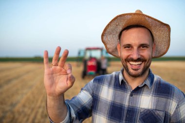 Ekose gömlekli ve hasır şapkalı yakışıklı çiftçinin portresi traktörün önünde tarlada işaret gösteriyor.