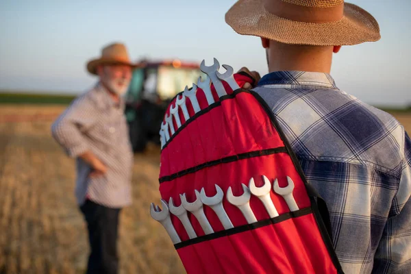 若い農家の肩にレンチのセットを保持し フィールド内の壊れたトラクターに歩くの背面ビュー — ストック写真