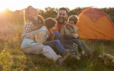 Dört kişilik bir aile bir tarlada bir arada oturur güneş batarken güler ve gülümserler. Kamp çadırlarının yanında keyifli bir anı paylaşıyorlar..
