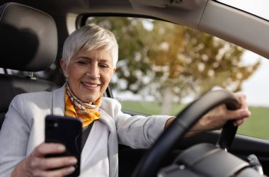 Neşeli yaşlı kadın araba sürerken elinde akıllı telefona bakıyor. Tehlikeli ve riskli bir aktivite. 