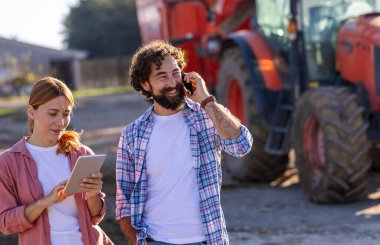 İki çiftlik işçisi dışarıda duruyor, traktörün önünde teknolojiyle uğraşıyorlar ve sığır çiftliğinde mikseri besliyorlar.