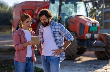 Dışarıda iki çiftlik işçisi, traktörün önünde tabletle uğraşıp sığır çiftliğinde mikserle besleniyor.