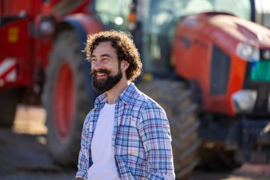 Kıvırcık saçlı ve sakallı genç çiftçinin portresi traktörün önünde duruyor sığır çiftliğinde yem karıştırıcıyla.